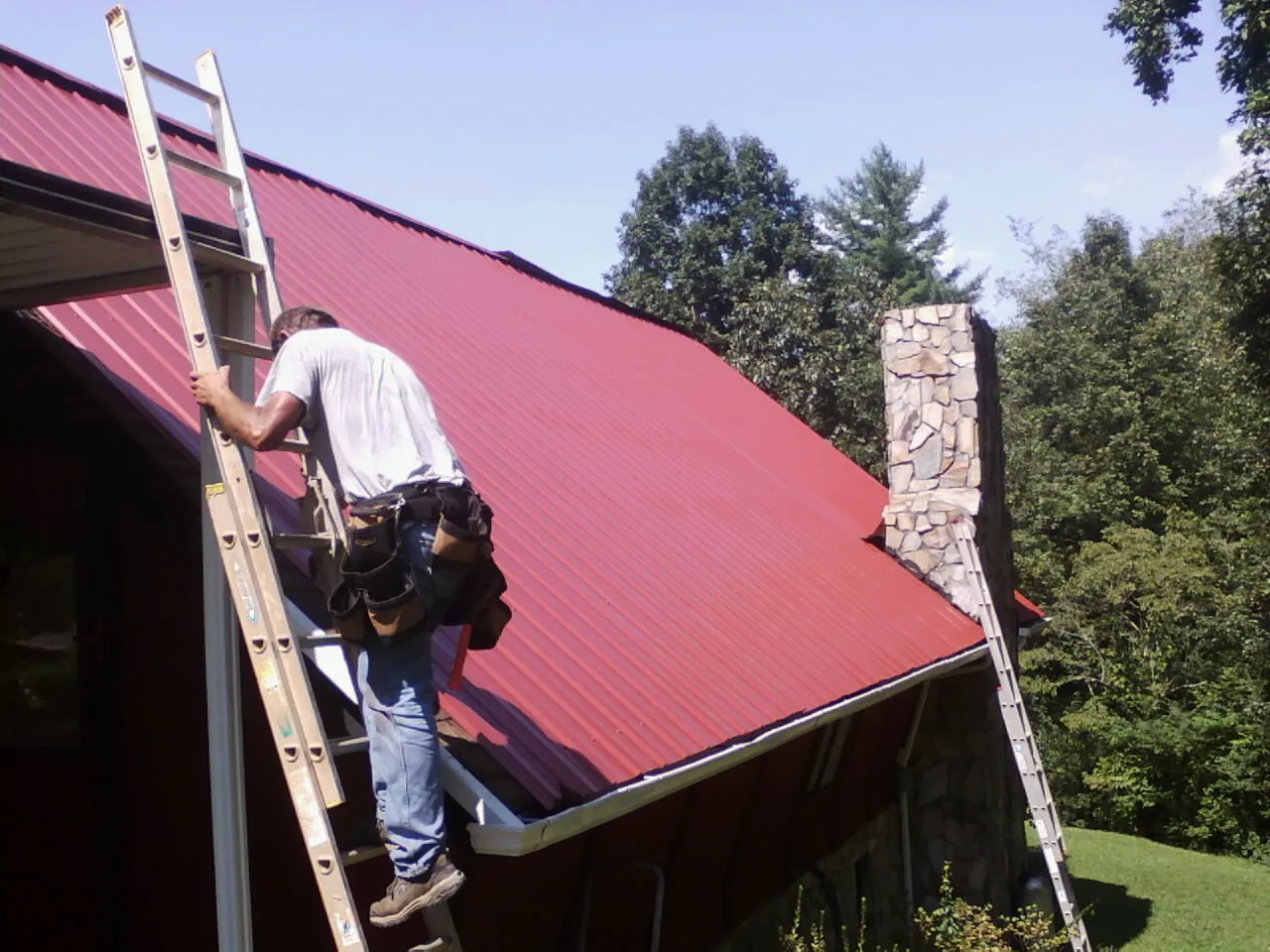 Roofer on a ladder beside a red metal roof and stone chimney — Soffit & Fascia Repair by Roofing Brothers in Sparks Glencoe
